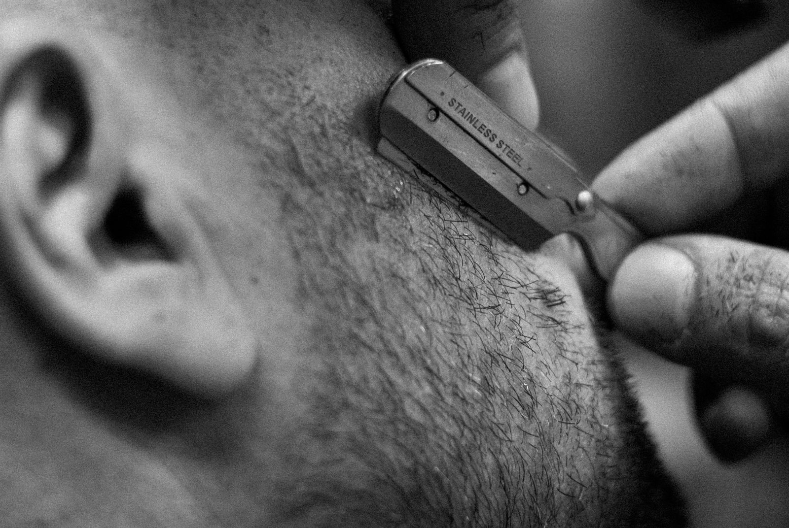 Black and white close-up of a straight razor edging the cheek line of a short, stubbly beard