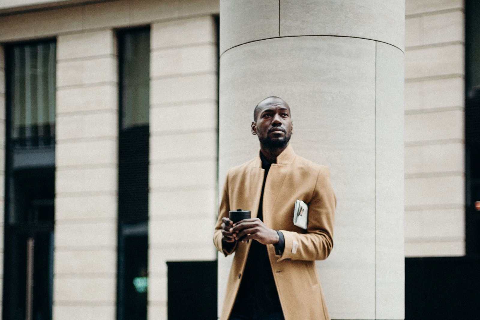 Man in a camel overcoat leaning against a stone column in the city, a classic look for men's coat trends fall 2026