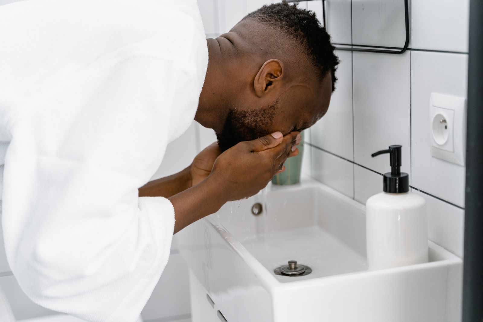 Bearded man in a white bathrobe leaning over a bathroom sink and splashing water on his face