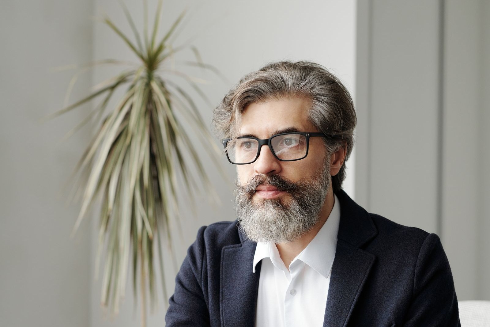 Grey-bearded man with glasses and short grey hair in a dark suit and white open-collar shirt, sitting in a bright modern office