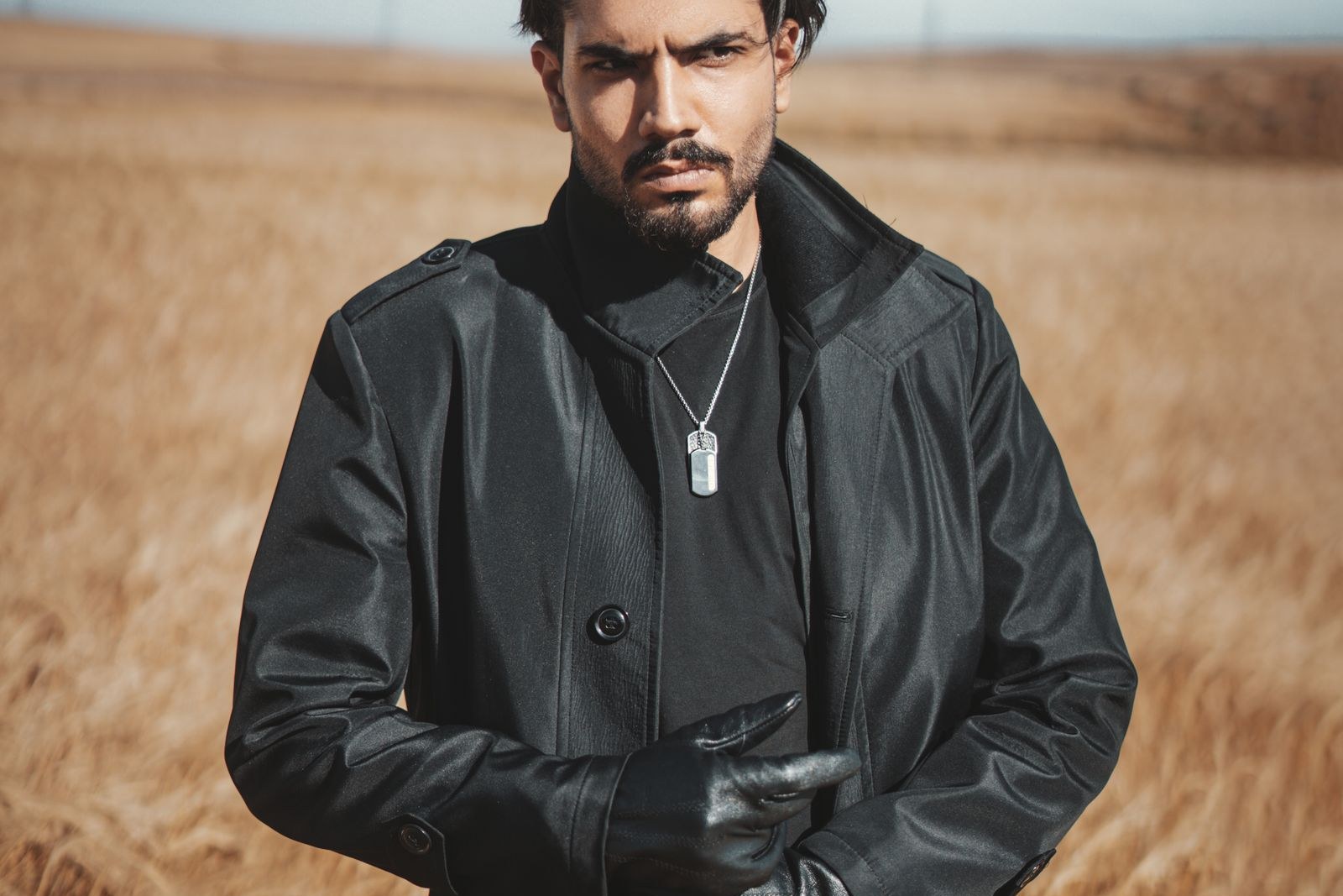 Bearded man with swept-back dark hair in a long black leather coat and black leather gloves, standing in a golden wheat field