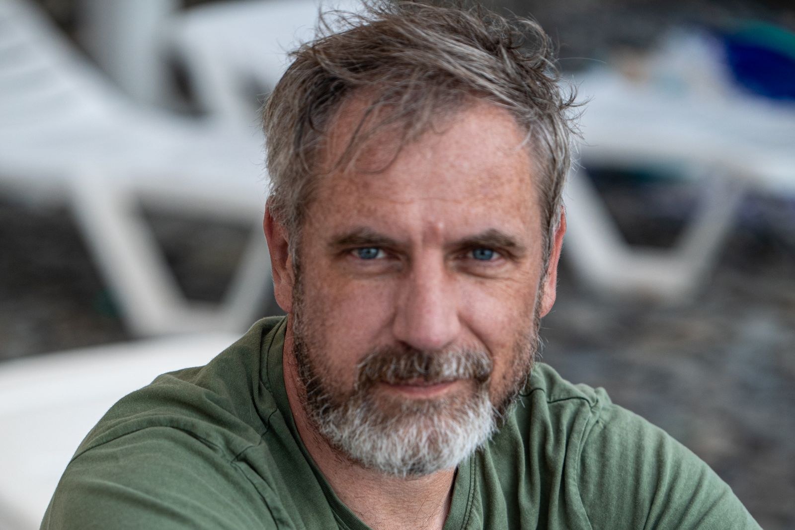 Middle-aged man with tousled grey hair and a short salt-and-pepper beard, wearing a green T-shirt and sitting outdoors