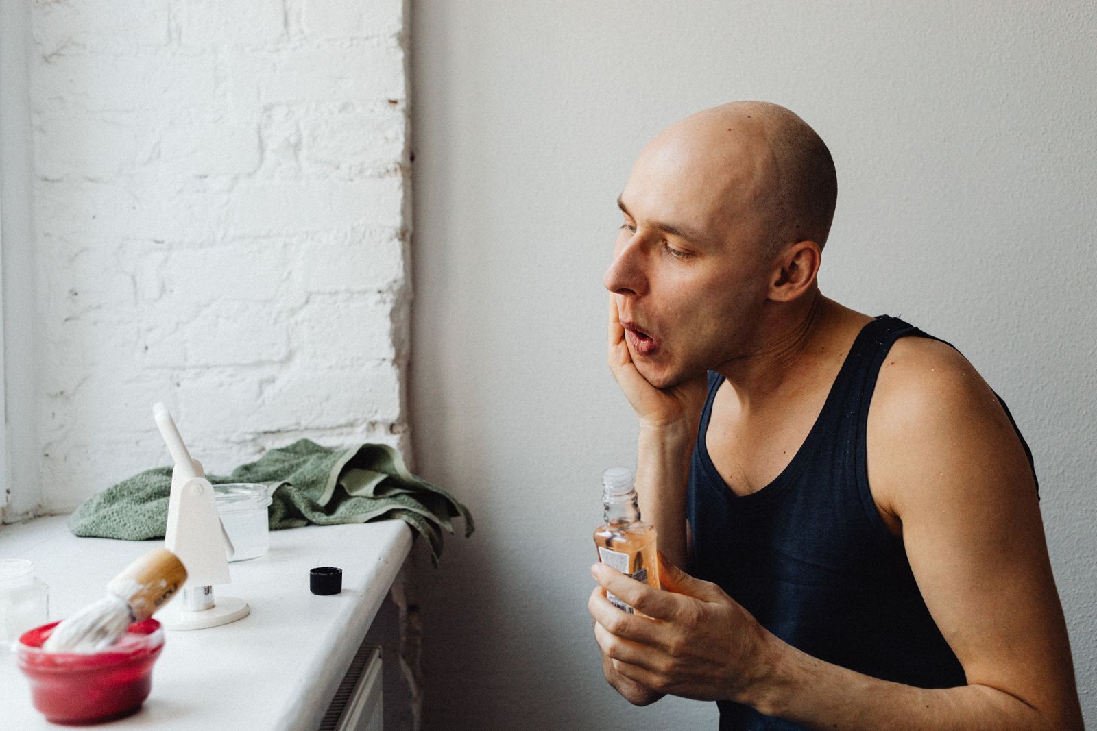 Bald, clean-shaven man in a black tank top touching his cheek and holding a bottle of aftershave by a bright window, with a shaving brush and red bowl on the sill
