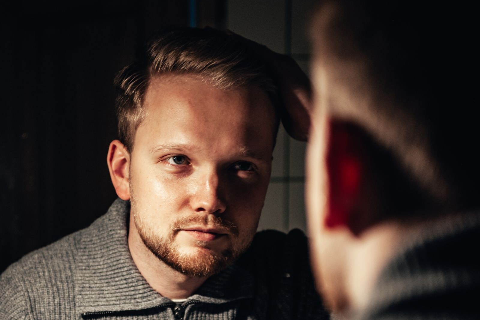 Bearded man with short brown hair touching the side of his head while looking into a mirror in a dim room