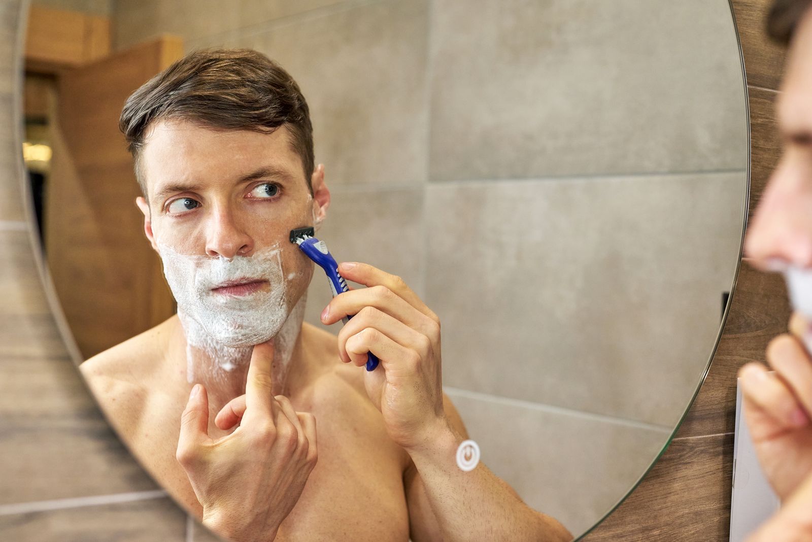 Young man with shaving foam on his chin and jaw shaving with a blue cartridge razor while looking into a round bathroom mirror