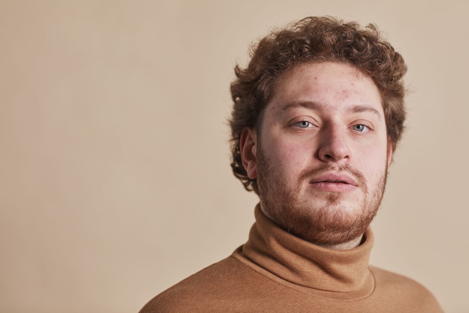 Bearded man in a camel turtleneck sweater against a warm beige backdrop, men's sweater trends fall 2026