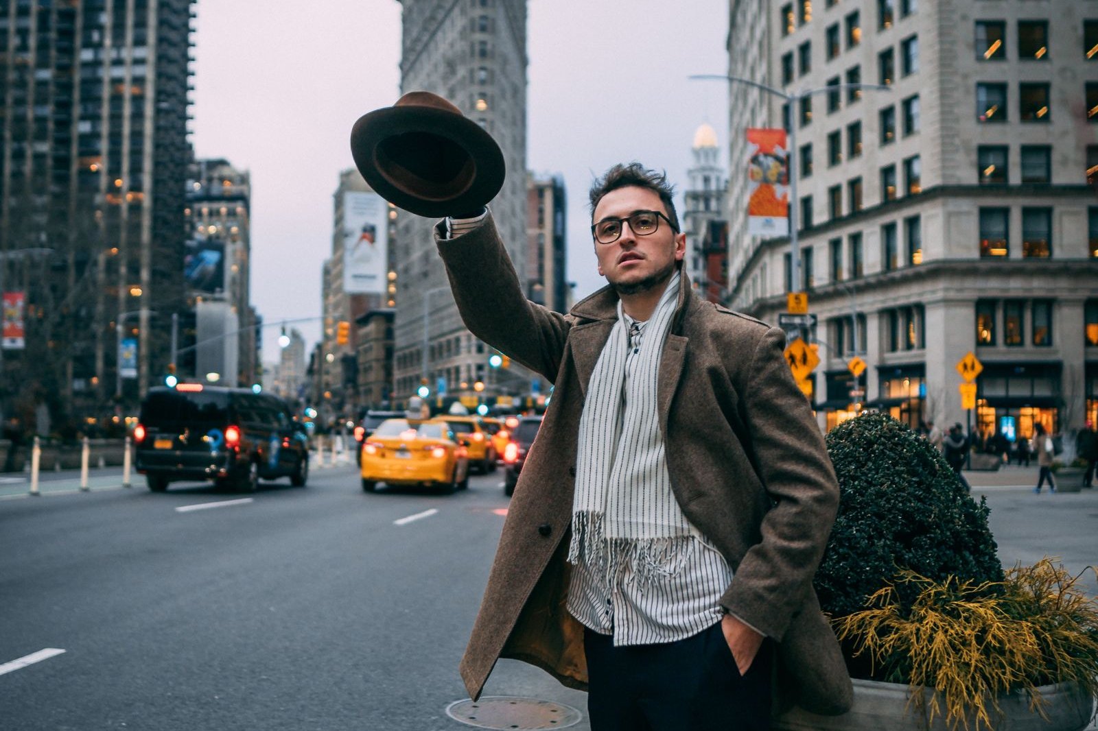 Man with glasses and short beard in a brown overcoat and striped scarf raising a brown felt fedora on a New York street, with the Flatiron Building behind him