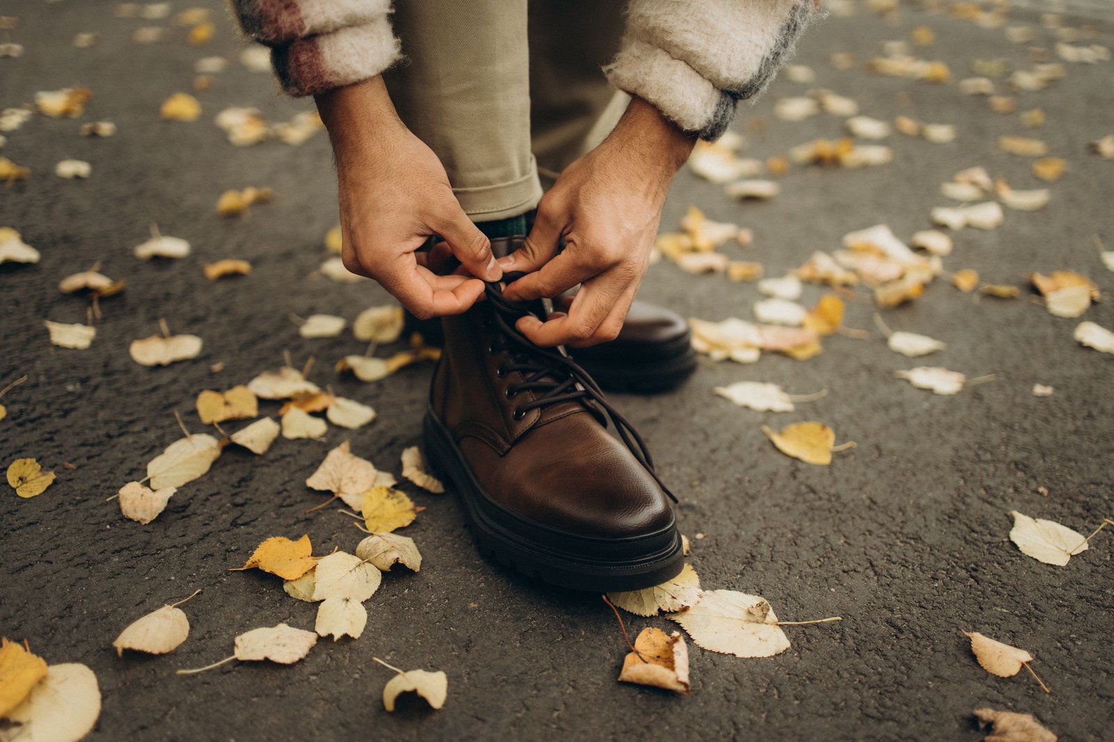 Men's boot trends fall 2026: a man in khaki chinos and a shearling-trimmed jacket tying the laces of his dark brown leather lace-up boots on an asphalt path scattered with autumn leaves