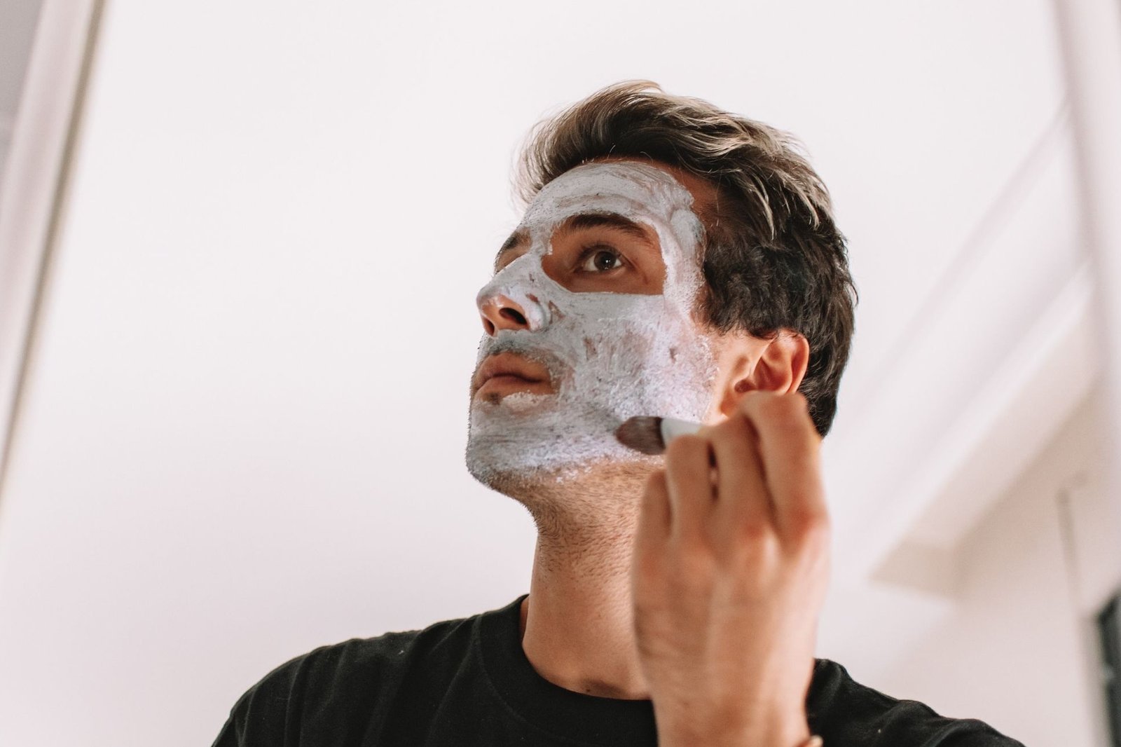 Fair-skinned man with tousled dark hair in a black T-shirt brushing a grey clay face mask onto his cheek in a bright room